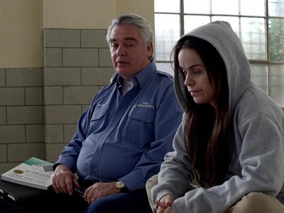 screen shot from orange is the new black season 2 episode 10 showing a male counselor talking with a female inmate wearing a hoodie in the group therapy room. There is a copy of the first edition of mind over mood siting on arm of the counselors chair.